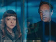A young white woman and an older man stand behind reflected glass looking serious