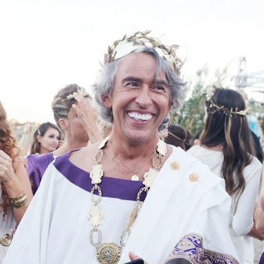 An older fake tanned man, smiling with bright white teeth, wearing ancient Greek fancy dress costume in a crowd of similarly dressed people