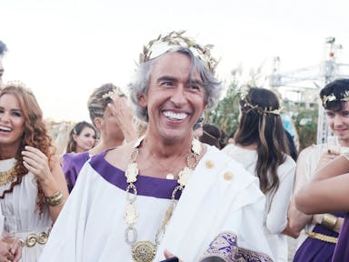 An older fake tanned man, smiling with bright white teeth, wearing ancient Greek fancy dress costume in a crowd of similarly dressed people