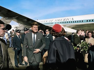 A white man in a suit, JFK, walking away from an airplane with 'United States of America' written on it, surrounded by official people