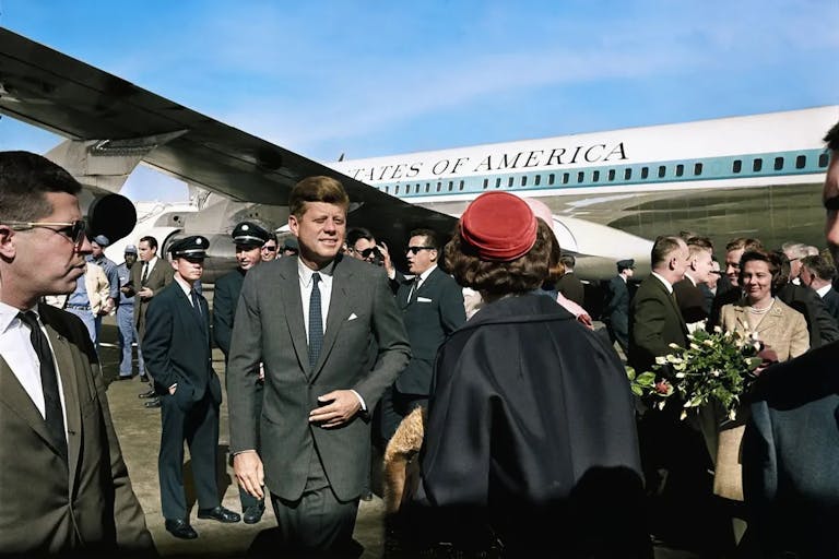 A white man in a suit, JFK, walking away from an airplane with 'United States of America' written on it, surrounded by official people