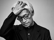 Black and white still of an older Japanese man with black rimmed glasses and dark clothing, looking down, running a hand through his hair