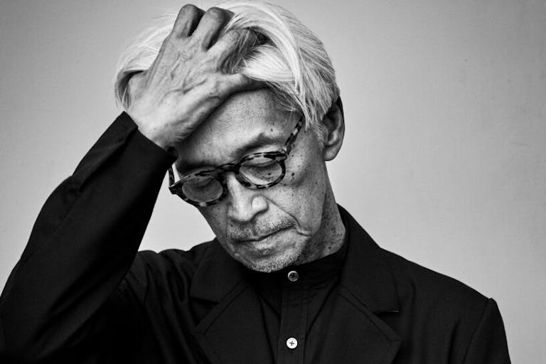 Black and white still of an older Japanese man with black rimmed glasses and dark clothing, looking down, running a hand through his hair