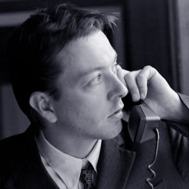 Black and white still of a white man in a suit holding up a landline phone to his ear while looking suspiciously out the window 