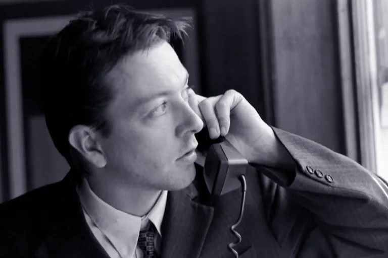 Black and white still of a white man in a suit holding up a landline phone to his ear while looking suspiciously out the window