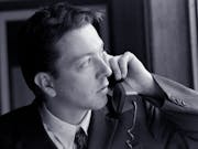 Black and white still of a white man in a suit holding up a landline phone to his ear while looking suspiciously out the window