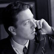 Black and white still of a white man in a suit holding up a landline phone to his ear while looking suspiciously out the window
