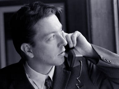 Black and white still of a white man in a suit holding up a landline phone to his ear while looking suspiciously out the window 