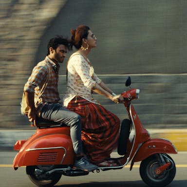A Pakistani trans woman riding a red moped wearing white and red salwar kameez and large good earrings, with a Pakistani man sitting on the moped behind her