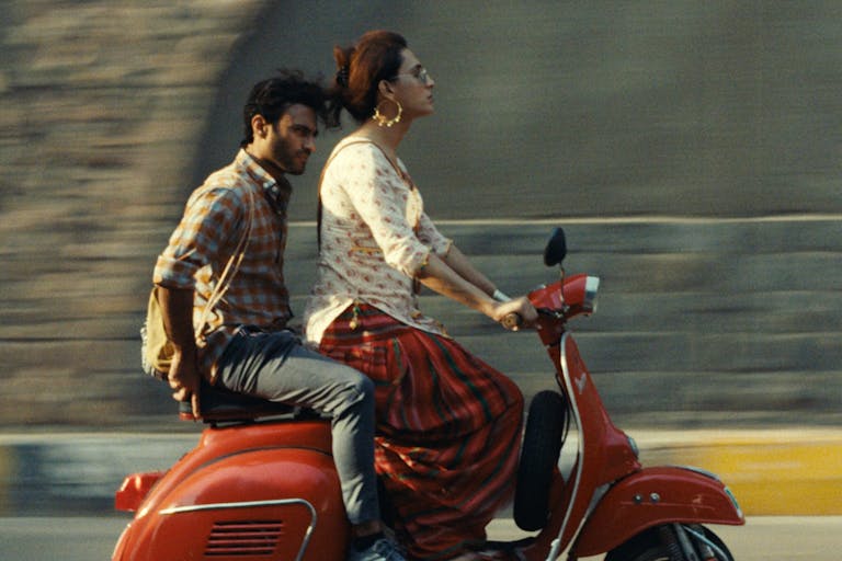 A Pakistani trans woman riding a red moped wearing white and red salwar kameez and large good earrings, with a Pakistani man sitting on the moped behind her
