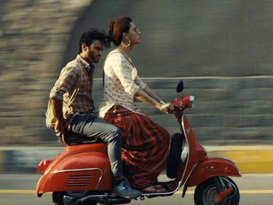 A Pakistani trans woman riding a red moped wearing white and red salwar kameez and large good earrings, with a Pakistani man sitting on the moped behind her