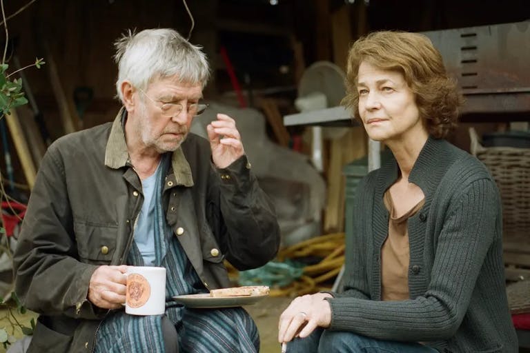An older white man and woman sit outside their shed drinking a cup of tea and eating a slice of toast