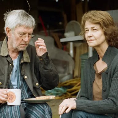 An older white man and woman sit outside their shed drinking a cup of tea and eating a slice of toast