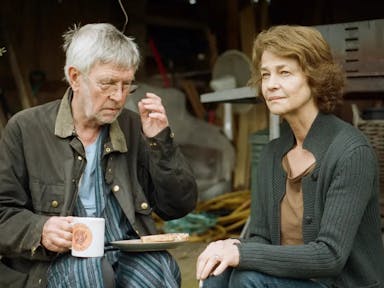 An older white man and woman sit outside their shed drinking a cup of tea and eating a slice of toast