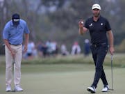 White male golfer in dark clothing holding a gold ball and golf club walking across a golf course