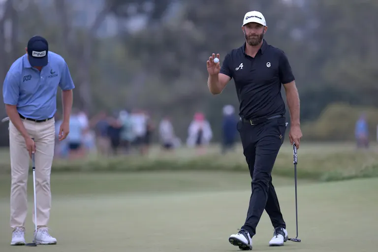 White male golfer in dark clothing holding a gold ball and golf club walking across a golf course