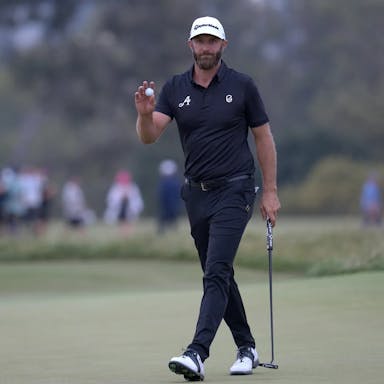 White male golfer in dark clothing holding a gold ball and golf club walking across a golf course