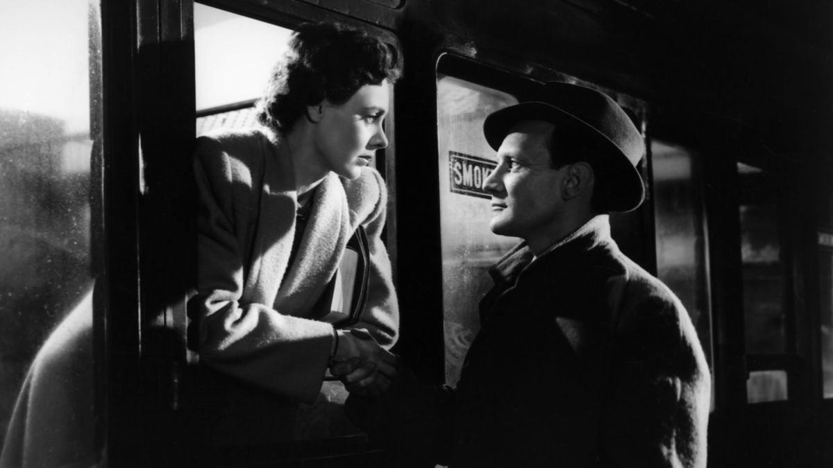A 1940s black-and-white picture of a woman leaning out of a train window, while a man stands outside. Both very close to eachother, looking at eachother romantically