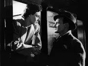 A 1940s black-and-white picture of a woman leaning out of a train window, while a man stands outside. Both very close to eachother, looking at eachother romantically