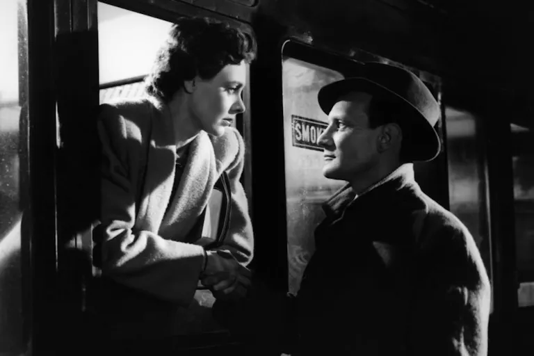A 1940s black-and-white picture of a woman leaning out of a train window, while a man stands outside. Both very close to eachother, looking at eachother romantically