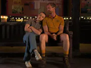 A young girl staring straight ahead, resting her head on the shoulder of an older man in a camp counsellor uniform