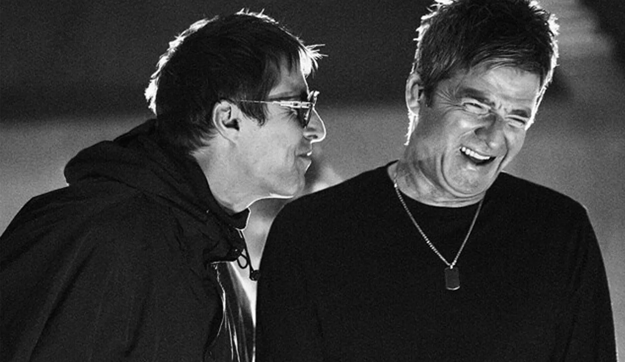 A black-and-white close-up still of two older men (Liam and Noel Gallagher of the band Oasis) mid-laugh