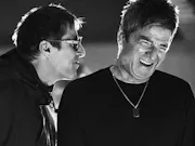 A black-and-white close-up still of two older men (Liam and Noel Gallagher of the band Oasis) mid-laugh