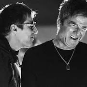 A black-and-white close-up still of two older men (Liam and Noel Gallagher of the band Oasis) mid-laugh