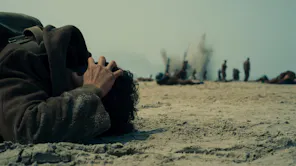 An army man laying on the floor face down with his hands over his ears, as explosions happen in the background