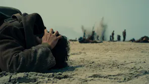 An army man laying on the floor face down with his hands over his ears, as explosions happen in the background