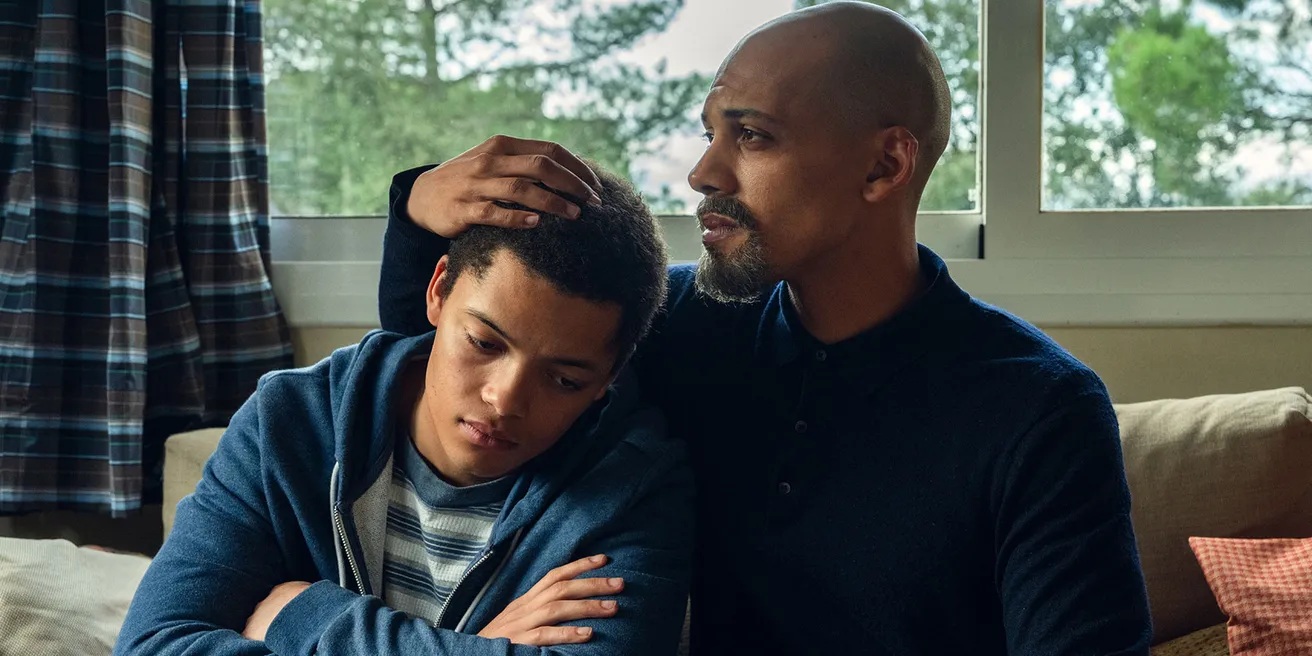 A black father and son sitting in a bedroom. The son, resting his head on the fathers shoulder
