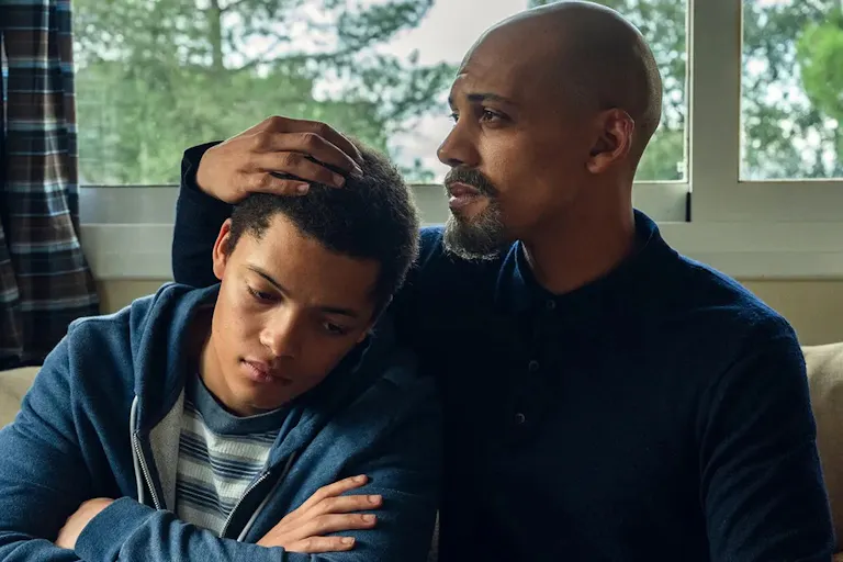 A black father and son sitting in a bedroom. The son, resting his head on the fathers shoulder
