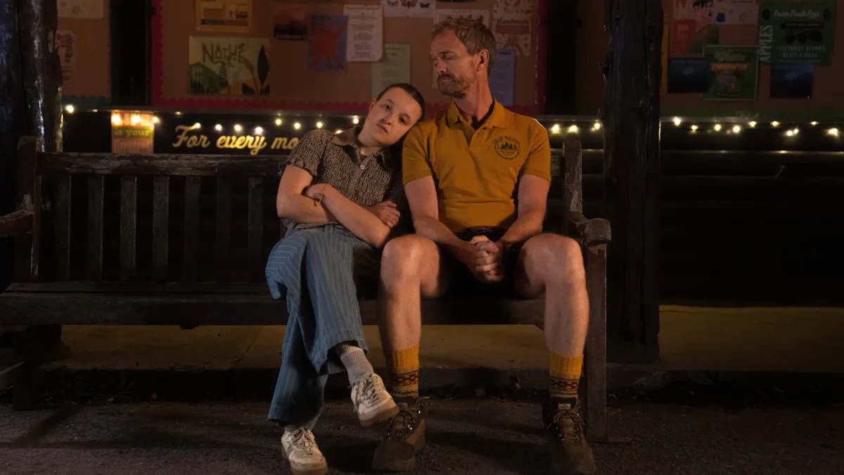 A young girl staring straight ahead, resting her head on the shoulder of an older man in a camp counsellor uniform