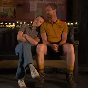 A young girl staring straight ahead, resting her head on the shoulder of an older man in a camp counsellor uniform