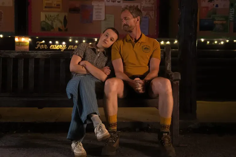 A young girl staring straight ahead, resting her head on the shoulder of an older man in a camp counsellor uniform
