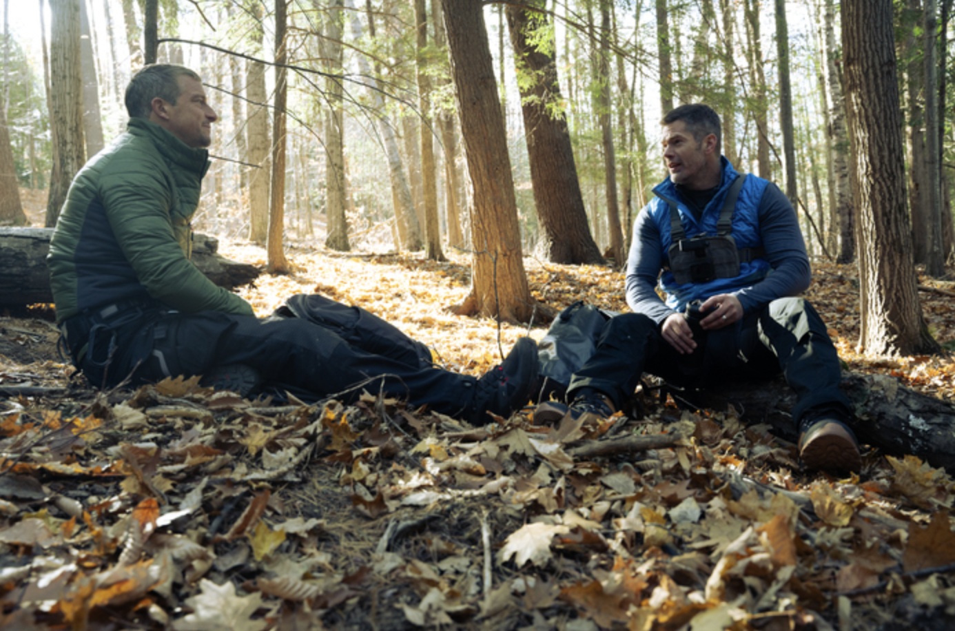 Two white men sitting on leaves in the middle of the woods, facing eachother