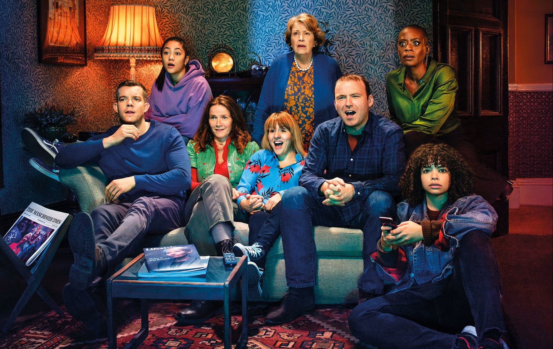 7 people of different ages and races siting around a couch with eyes glued to a TV, each wearing different colours and with a shocked look on their face