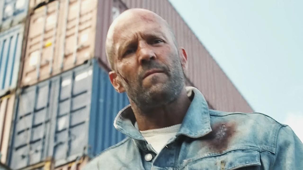 A bald white man looking down at the camera with an angry face, behind him some stacked shipping containers