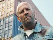 A bald white man looking down at the camera with an angry face, behind him some stacked shipping containers