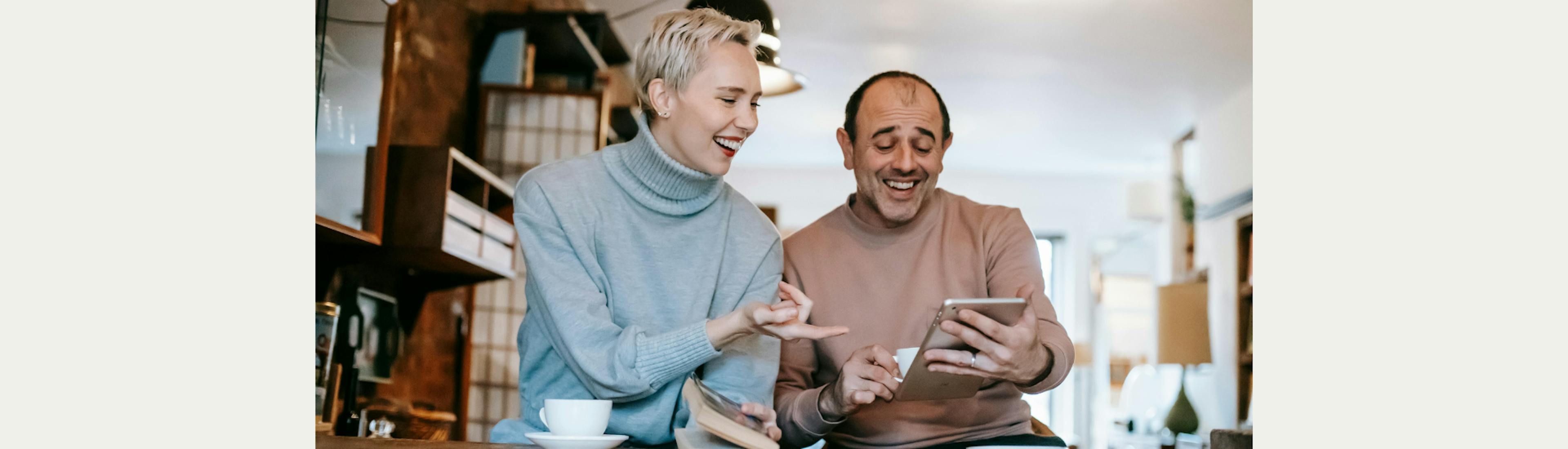 Een man en vrouw zitten in een cafe. Ze kijken lachend naar een tablet. 
