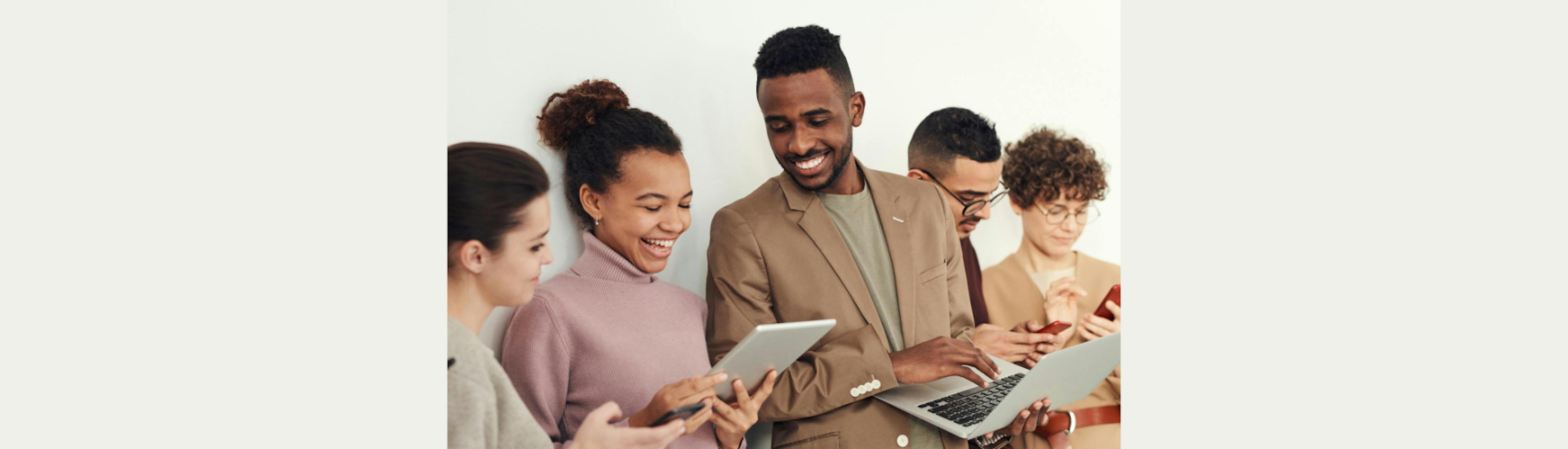 Een groep mensen staat op een rij, ze kijken vrolijk naar verschillende digitale schermen: een tablet, laptop en telefoon. 