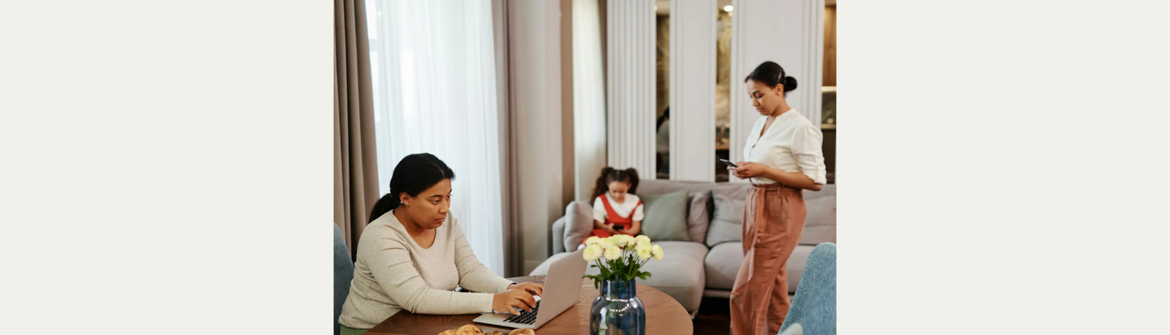 A woman is sitting at a dining table behind a laptop, a younger woman is standing with a phone in her hands, and a girl is sitting on the couch with a phone in her hands. They all look dejected.
