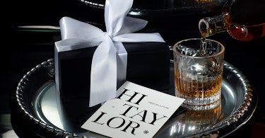 Image of customer appreciation gifts from Sculpture: a black box with a white satin bow, a fancy glass with whisky being poured into it, and a white card on a black tray.