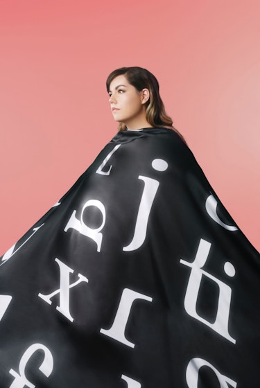 Nadège Dionne-Tremblay, co-founder of Sculpture, in front of a pink-coral background, draped in a long black cape featuring Sculpture the font, looking away.