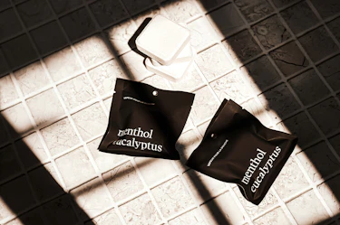 Two Marée Menthol Eucalyptus shower steamers in matte black packaging displayed on a tiled surface beside unwrapped shower steamers in natural light.