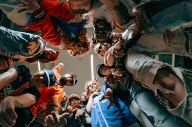 A group of people gathered in a circle, seen from below, look toward the camera. They wear colorful sporty and streetwear outfits. The scene takes place indoors and reflects the collective energy of OFFSTAGE Club Sportif.