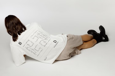 A person lying on their side in a studio against a light background, wearing a white t-shirt, beige shorts, and black boots. The back of the t-shirt features a graphic layout with linear drawings and the text “BALLET,” “DIVERTIMENTO,” along with the letters “B” and “D,” arranged in a composition inspired by Ballet Divertimento’s graphic system.