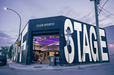 The exterior of a black building located on a city street corner, home to OFFSTAGE Club Sportif. The left facade displays the word “OFF” and the right facade displays the word “STAGE” in very large white letters. The entrance is open, revealing an interior lit with purple lighting.