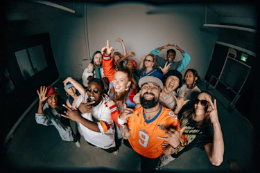 A group of people gathered indoors look toward the camera, posing expressively and smiling. They wear colorful sporty and streetwear outfits. The scene takes place in a minimal indoor space and highlights the collective spirit of OFFSTAGE Club Sportif.
