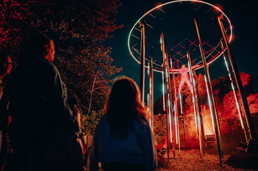 Immersive nighttime installation featuring a glowing human figure suspended at the center of a circular illuminated structure, surrounded by vertical light poles and red mist. Visitors watch from a forest path, within the Forest of Shadows experience by Moment Factory.